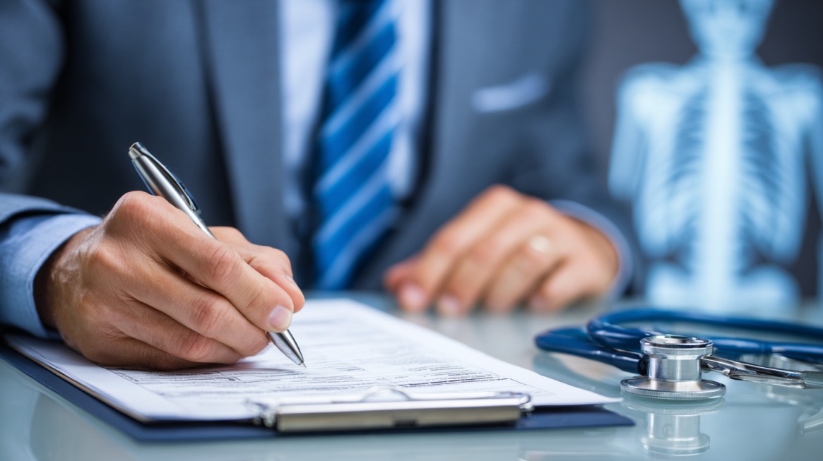 Close-up of a person in a suit signing insurance documents with a stethoscope and medical chart nearby